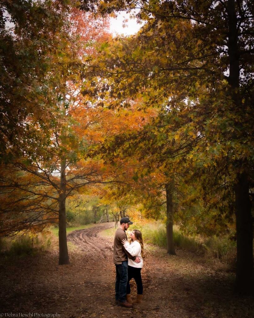 A couple embraces on a forest path surrounded by vibrant autumn foliage. The scene is peaceful and romantic, with soft light filtering through the trees.