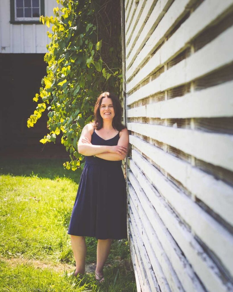 Smiling woman in a navy dress leans against a white wooden wall, arms crossed. Bright sunlight illuminates green vines and grass, creating a cheerful, serene atmosphere.