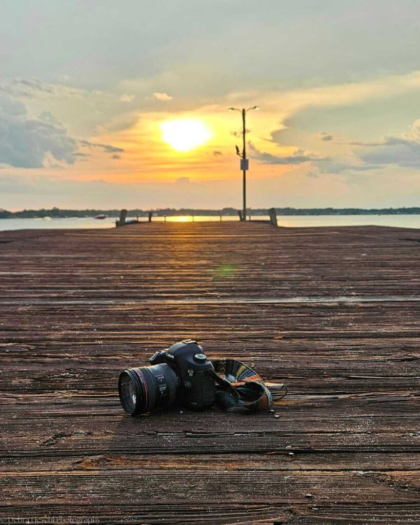 A DSLR camera sits on a wooden dock during a serene sunset, with the sun setting over calm waters in the background, creating a peaceful ambiance.