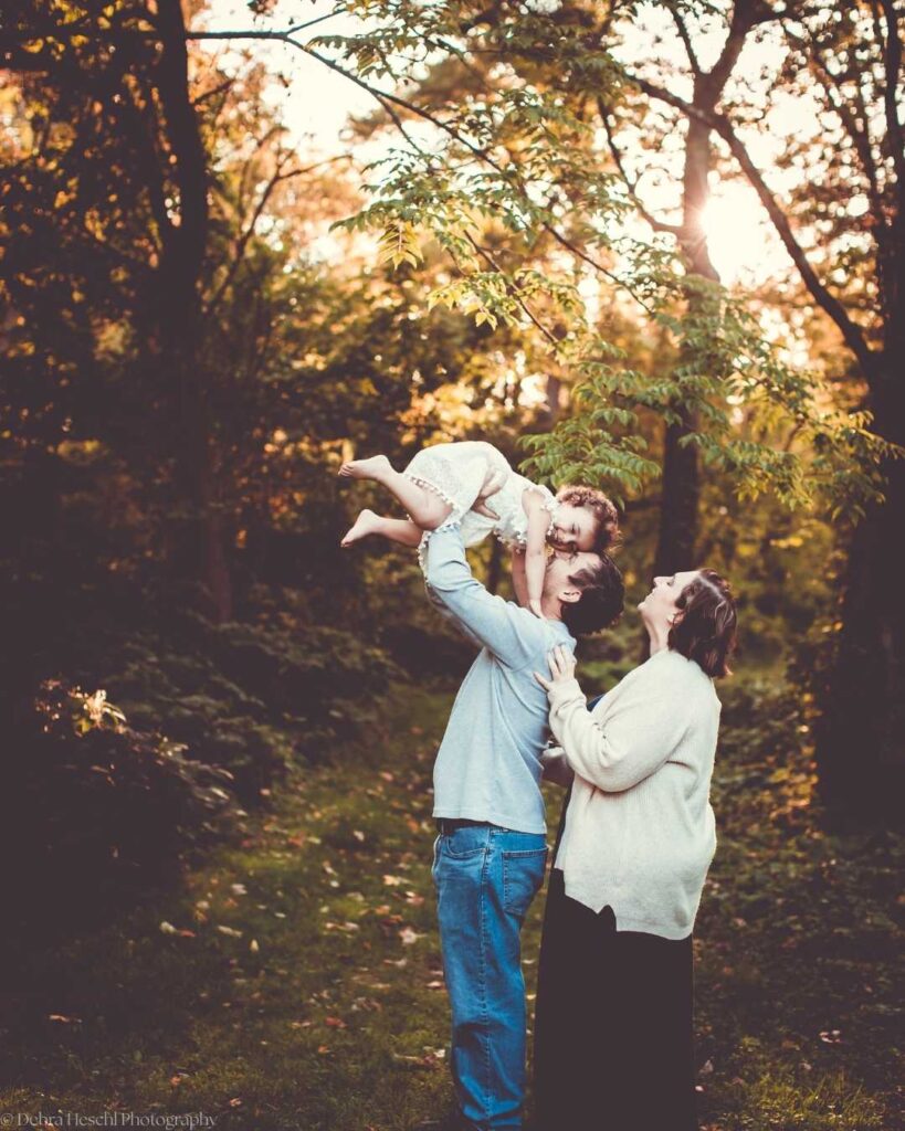 A joyful couple stands in a sunlit forest, the man lifting a smiling child overhead. The warm, glowing light creates a serene and happy atmosphere.