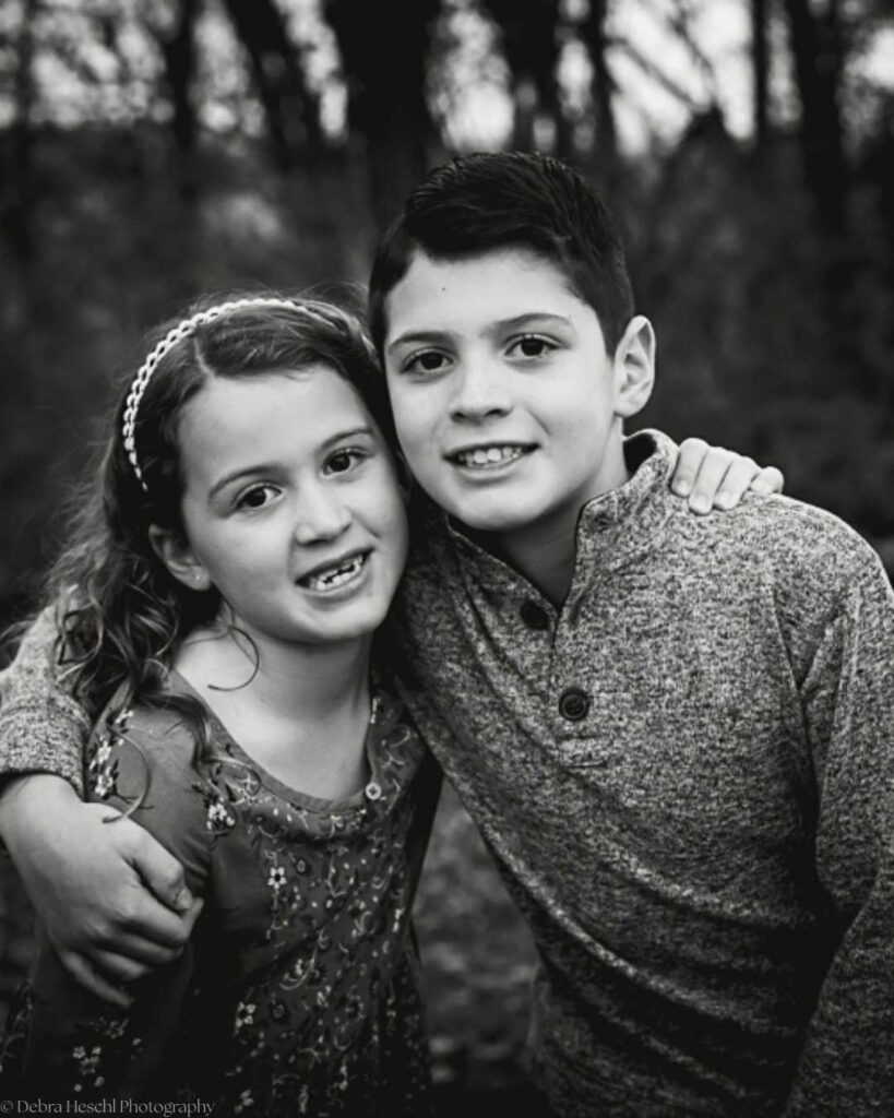 Black and white photo of a young boy and girl smiling with their arms around each other, set against a blurred forest backdrop, conveying warmth and friendship.
