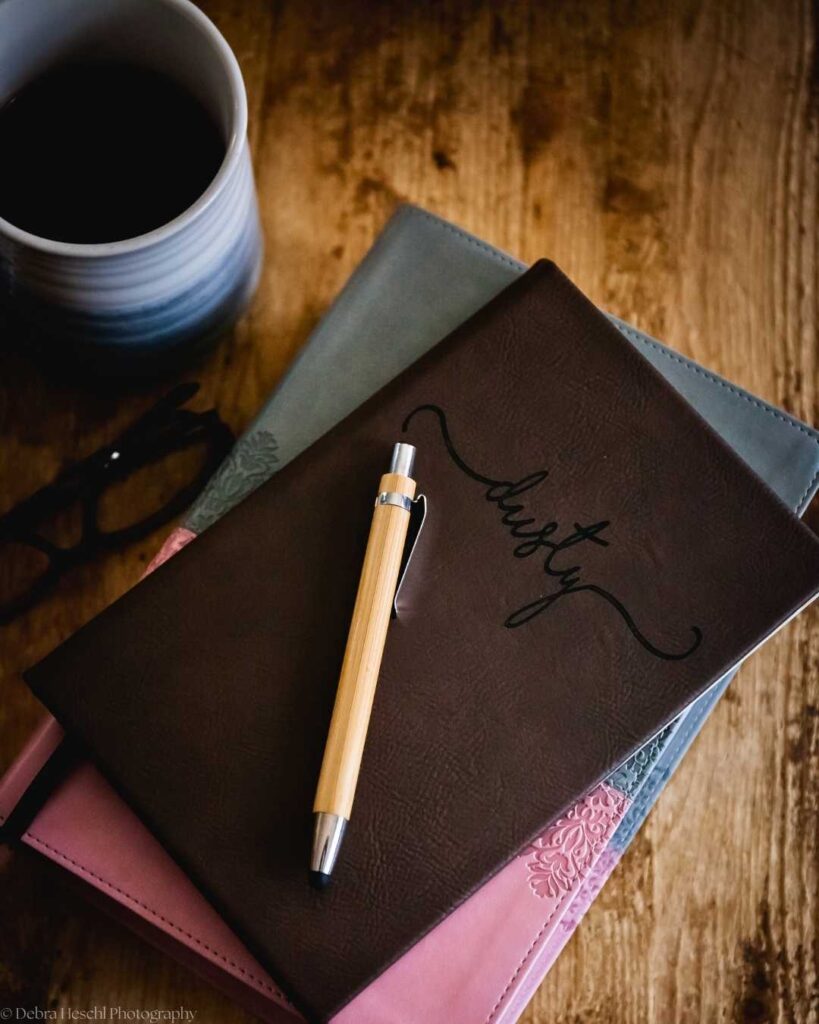 A wooden table with a cup of coffee, stacked journals in blue and pink, a brown journal with "dusty" in cursive, a pen, and glasses nearby.