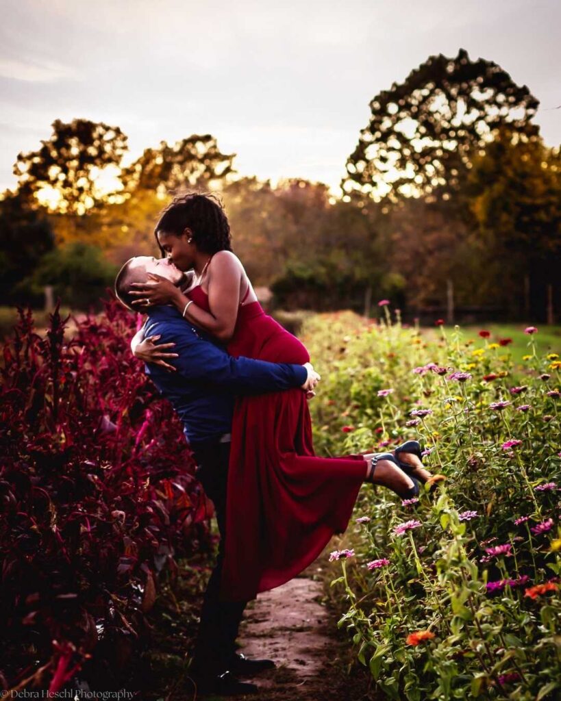 A couple embraces in a flower-filled garden at sunset. The woman, in a flowing red dress, is joyfully lifted by a man, creating a romantic scene.