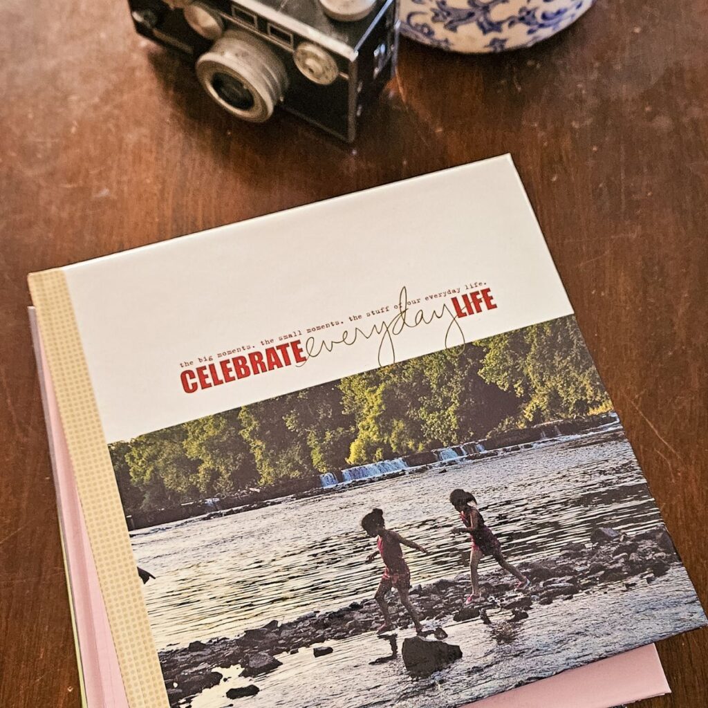 A book with "Celebrate Everyday Life" on the cover lies on a wooden table. Behind it is a vintage camera and a blue-patterned vase, suggesting nostalgia.