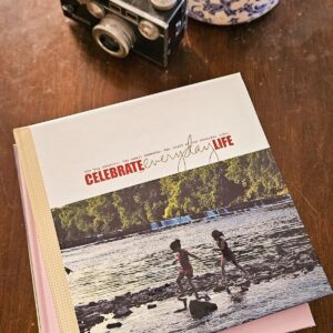 A book with "Celebrate Everyday Life" on the cover lies on a wooden table. Behind it is a vintage camera and a blue-patterned vase, suggesting nostalgia.