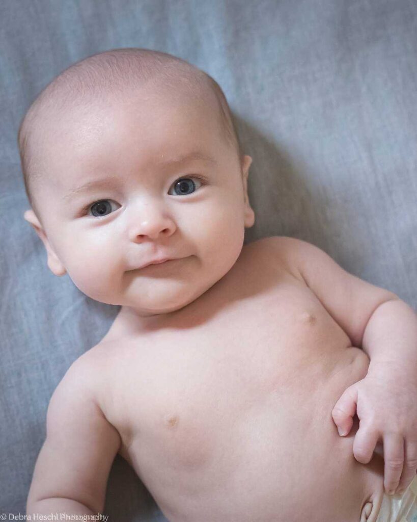 A smiling baby lies on a soft gray blanket, gazing upwards with bright eyes. The atmosphere is calm and serene, conveying warmth and innocence.