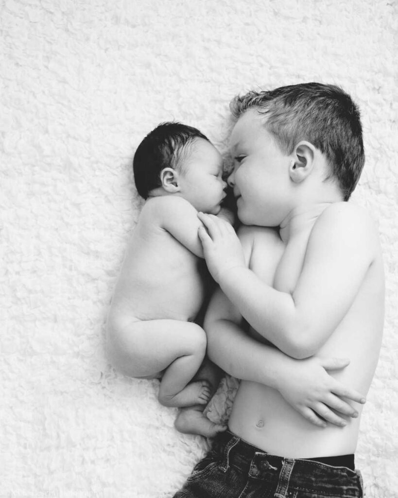 A black and white photo of a young boy gently hugging a newborn baby on a soft white blanket. The scene exudes warmth, love, and tenderness.