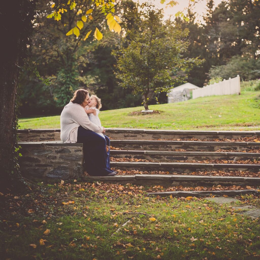 A woman and child share a warm embrace on stone steps surrounded by autumn leaves. The background shows a green field and trees under a soft, golden light.