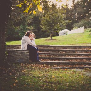 A woman and child share a warm embrace on stone steps surrounded by autumn leaves. The background shows a green field and trees under a soft, golden light.