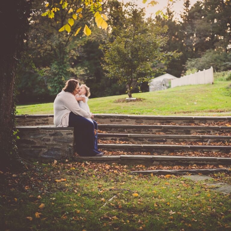 A woman and child share a warm embrace on stone steps surrounded by autumn leaves. The background shows a green field and trees under a soft, golden light.