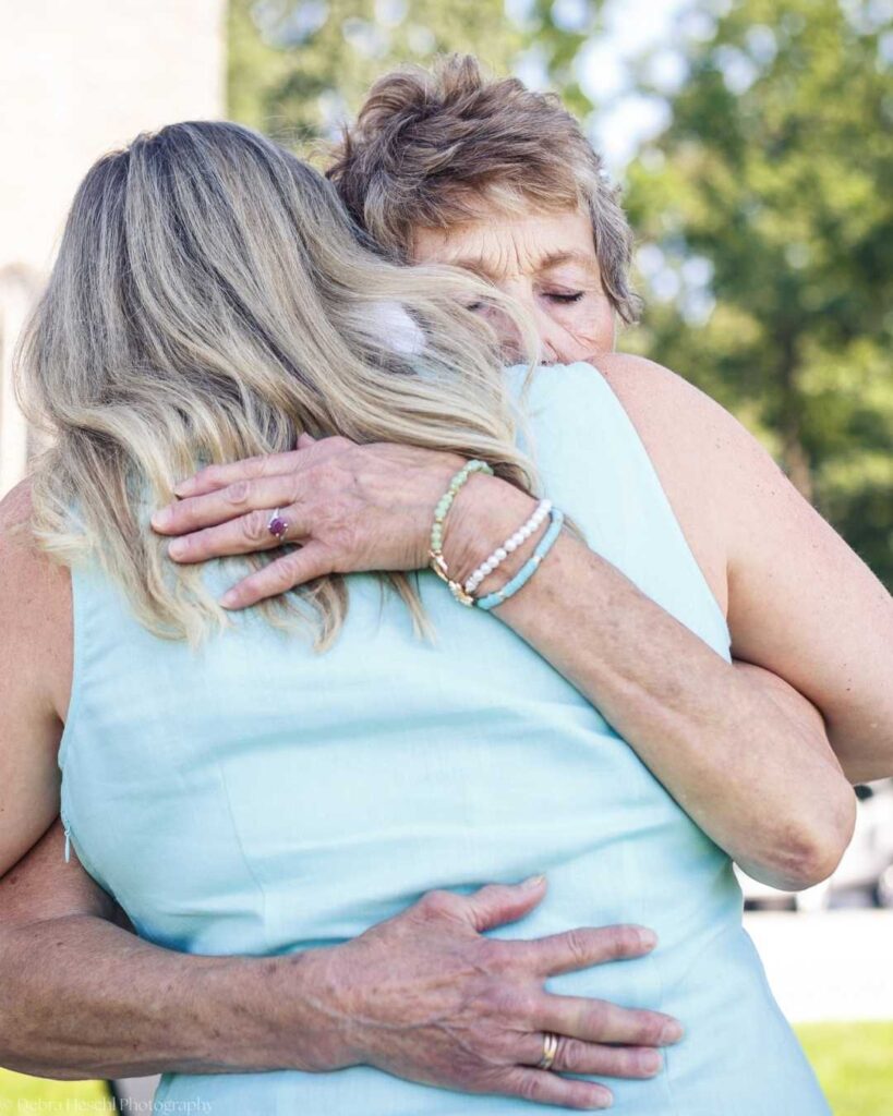 A loving embrace between two women outdoors, both wearing light blue clothing. One woman's face shows emotion, emphasizing warmth and connection.