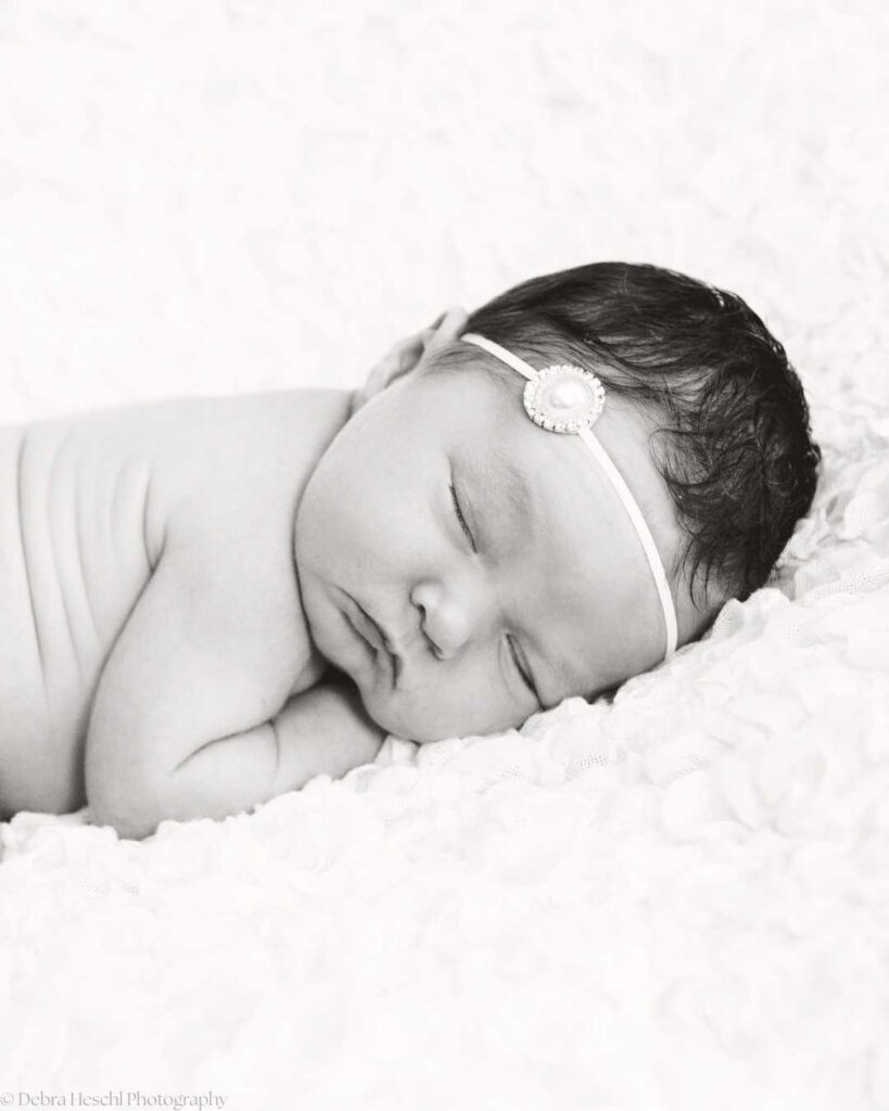 A peaceful black-and-white photo of a sleeping newborn with a headband adorned with a flower, lying on a soft, textured blanket.