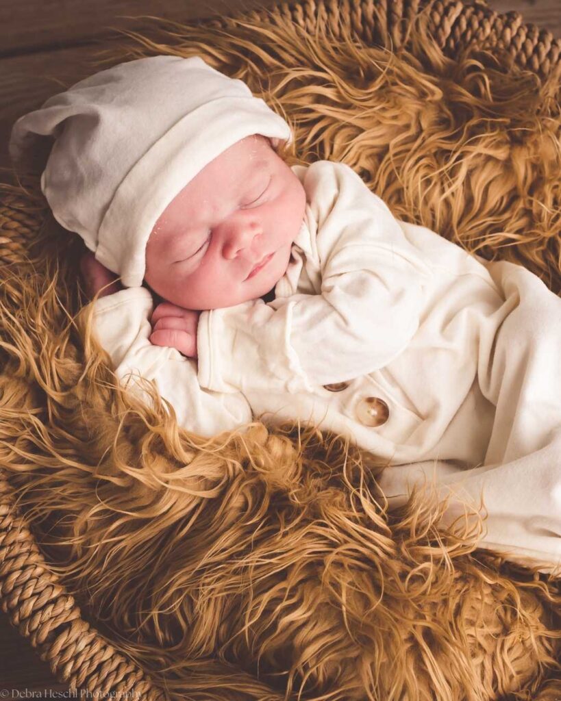 A newborn baby sleeps peacefully in a cozy, woven basket, wearing a soft, white onesie and matching hat, lying on a fluffy, golden-brown fur blanket.