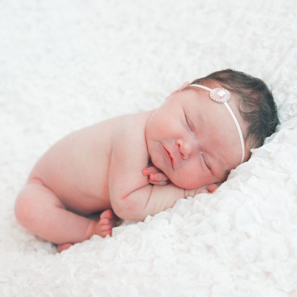 Newborn baby peacefully sleeping on a soft white blanket, wearing a delicate pink headband with a pearl. The scene conveys warmth and tranquility.