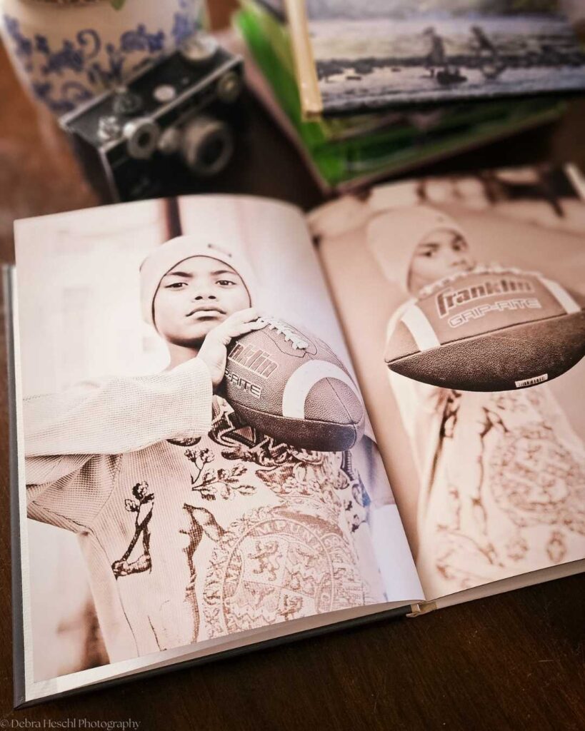 Black and white photo of a child holding a football, open on a table. Vintage camera, floral vase, and a pile of books are in the background. Calm and nostalgic.