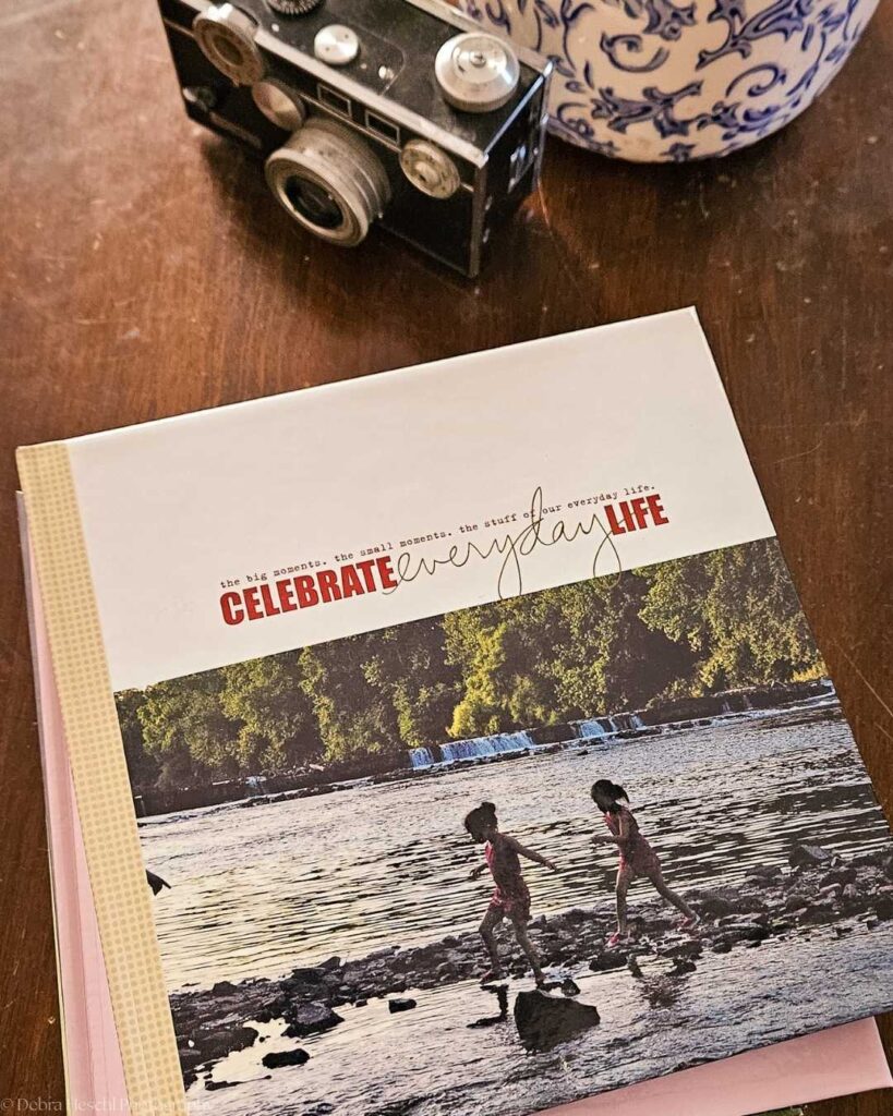 A vintage camera sits beside a floral-patterned vase on a wooden table. A book titled "Celebrate Everyday Life" features children playing by a river.