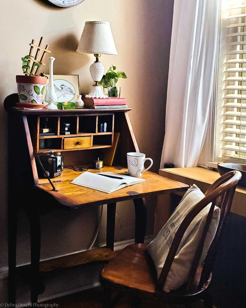 Cozy home office with a rustic wooden desk adorned with books, plants, a lamp, and framed art. A mug, notepad, and quill sit atop, under warm light.