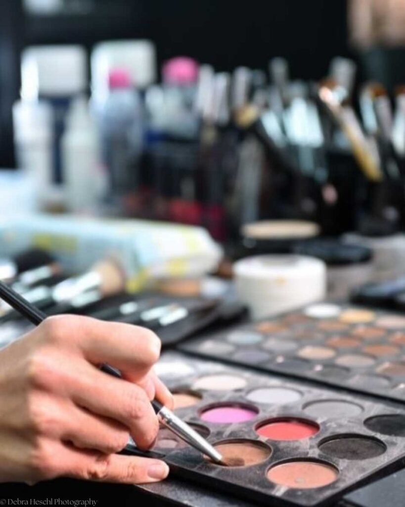 A hand holding a brush selects brown eyeshadow from a palette, with blurred makeup tools and products in the background, creating a professional and artistic atmosphere.