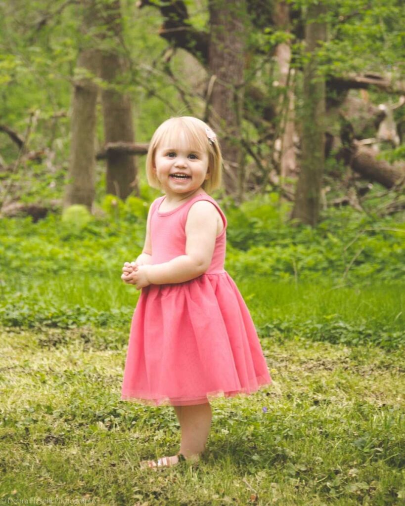 Toddler girl in pink dress laughing during outdoor spring photo session in the woods.