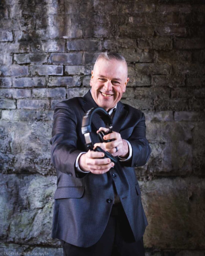 A smiling man in a suit playfully holds headphones towards the camera against a textured brick wall, creating a friendly and lively atmosphere.
