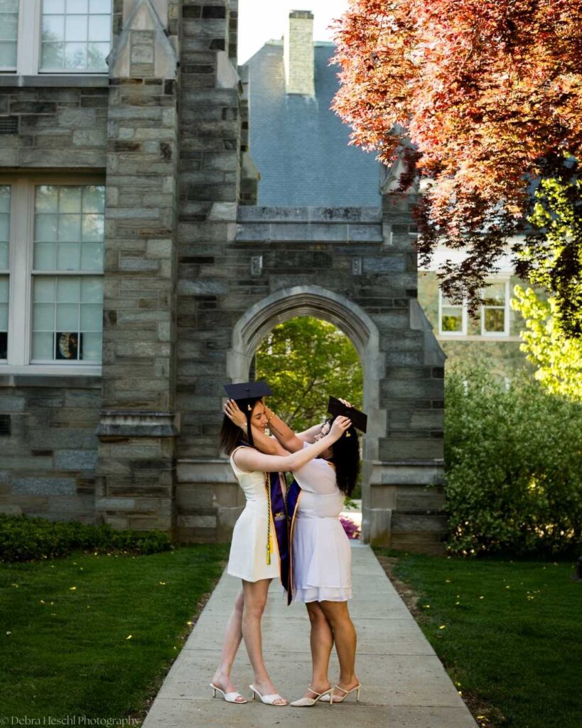 Best friends Leah and Miranda adjust each other's graduation caps in front of West Chester University's iconic stone archway.