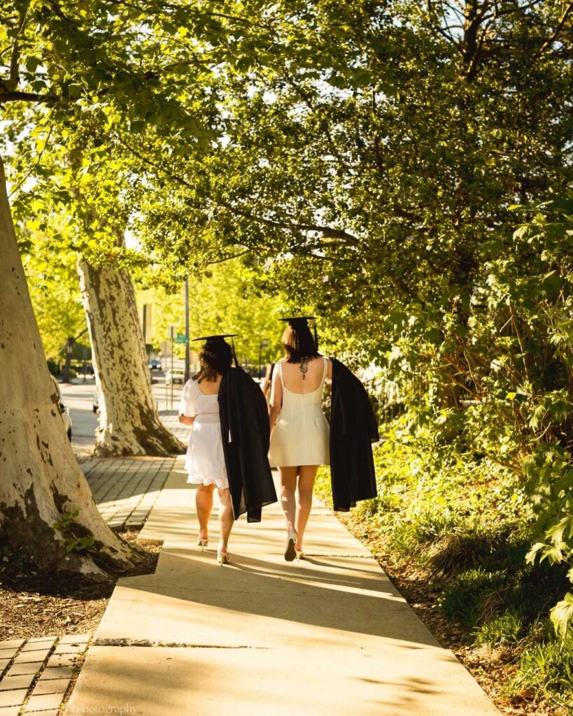 Best friends Leah and Miranda walk together beneath shaded trees in their graduation gowns at West Chester University