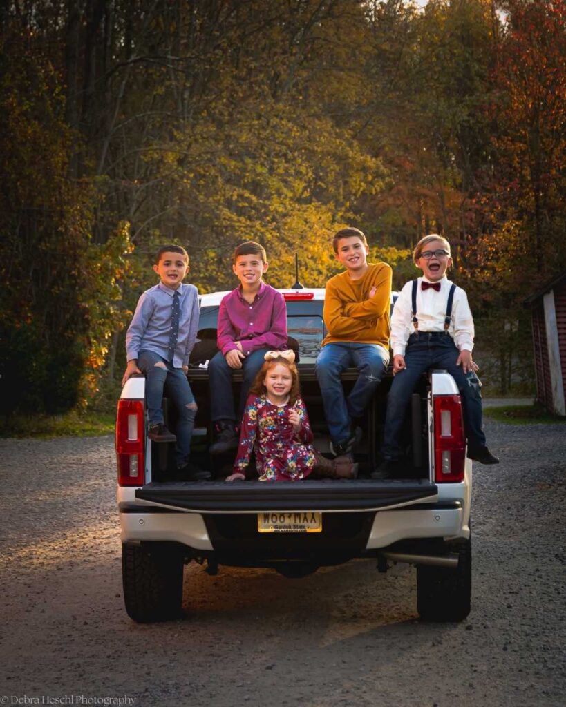 Candid group photo of five children sitting and laughing on a truck bed surrounded by golden hour fall foliage