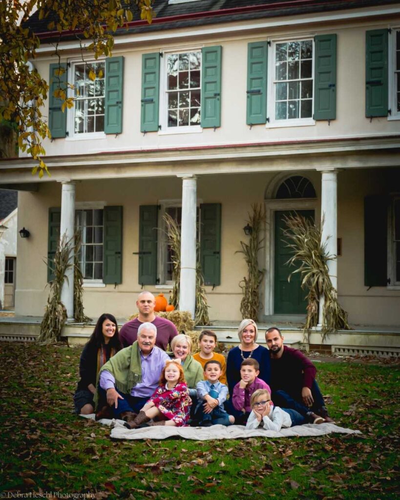 A large family poses cheerfully in front of a two-story house with green shutters. Autumn decorations, including pumpkins and corn stalks, add warmth.