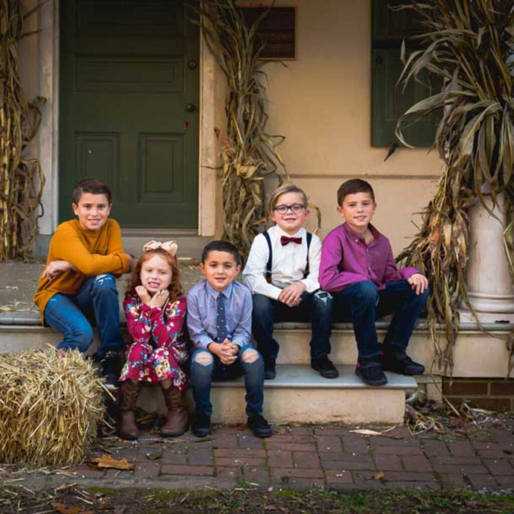 Five children sit on porch steps with corn stalks in the background. They're smiling, dressed in colorful, autumnal clothing, creating a warm, festive atmosphere.
