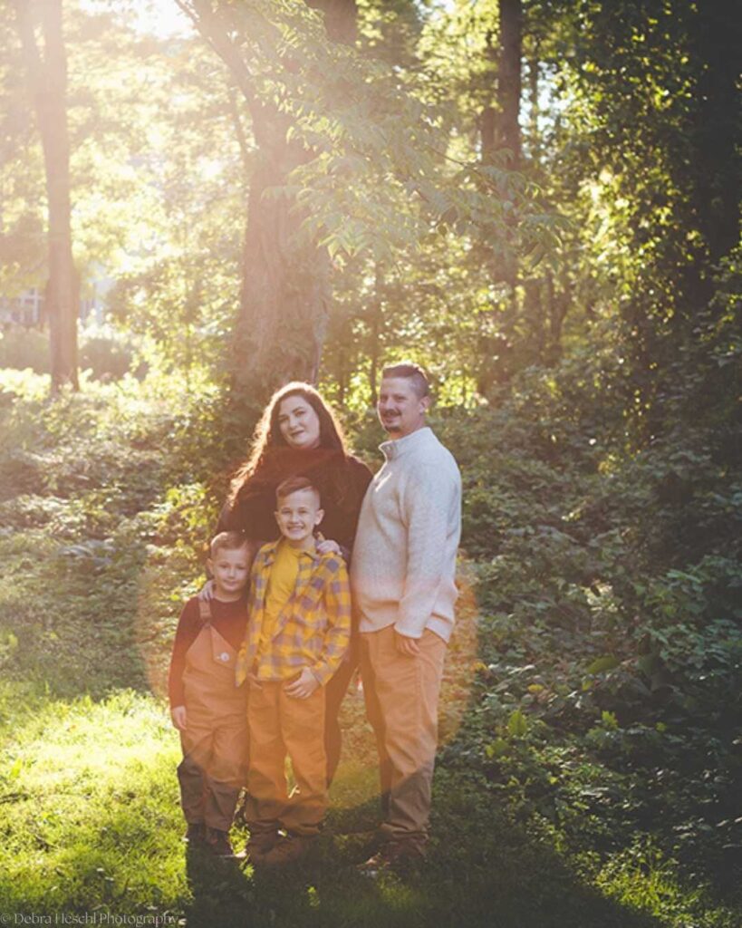 Family of four smiling together in a sunlit wooded setting during an outdoor family photography session with Debra Heschl Photography