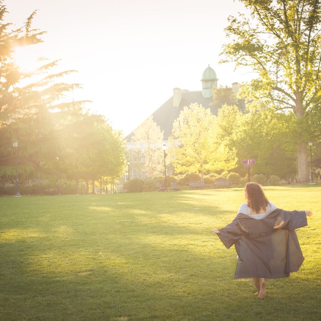 A woman in a graduation gown walks barefoot on a sunlit lawn surrounded by trees, with a historic building in the background, evoking a sense of joy and achievement.