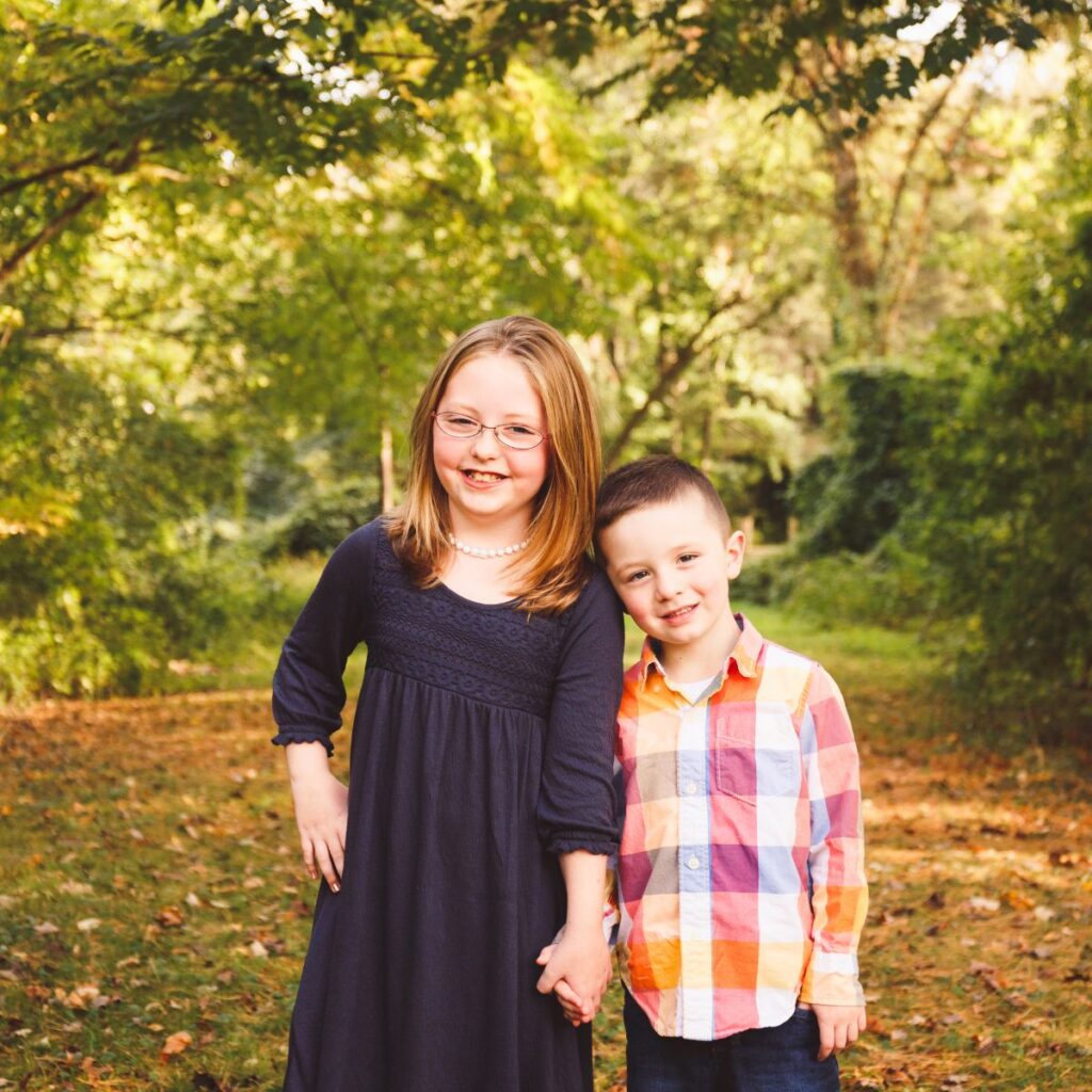 Natural sibling portrait of a brother and sister holding hands and smiling outdoors in fall foliage.