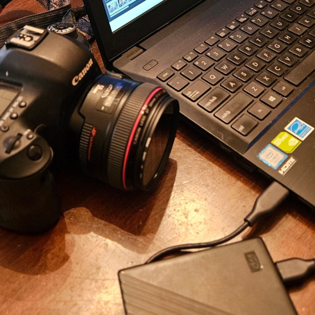 Backing up and editing photos after your session: Canon camera lens beside laptop and external hard drive.