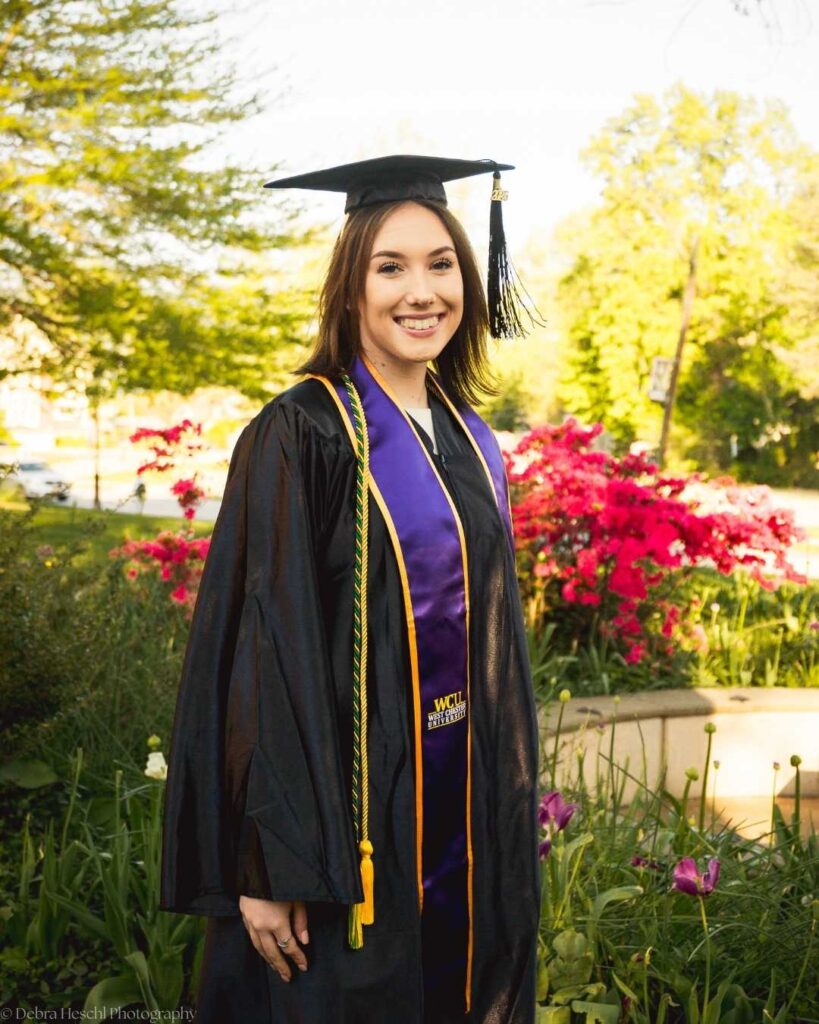 Smiling West Chester University graduate Leah in cap and gown with purple and gold honor cords among spring flowers.
