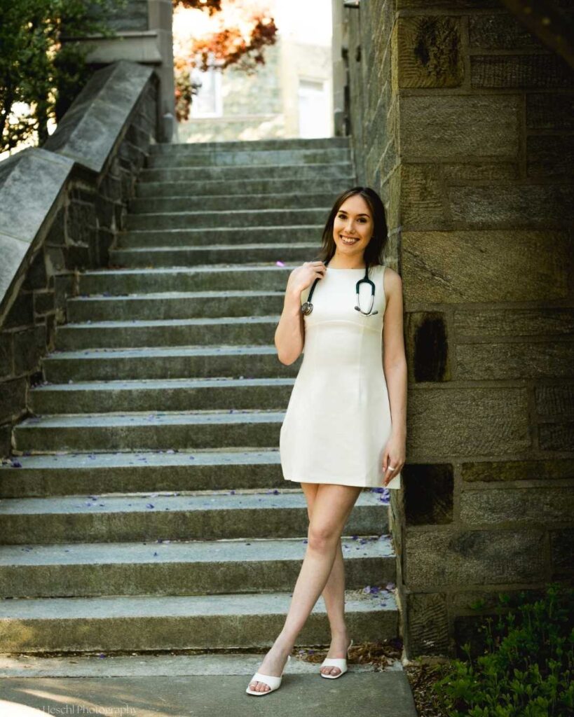 Respiratory Therapy graduate Leah poses on stone stairs in a white dress with stethoscope at West Chester University, captured by Debra Heschl Photography.