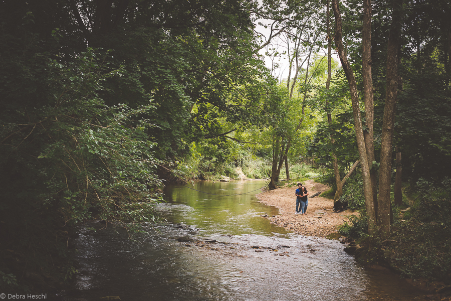 A couple stands on a tranquil forest riverbank, surrounded by lush green trees, creating a serene and intimate atmosphere.