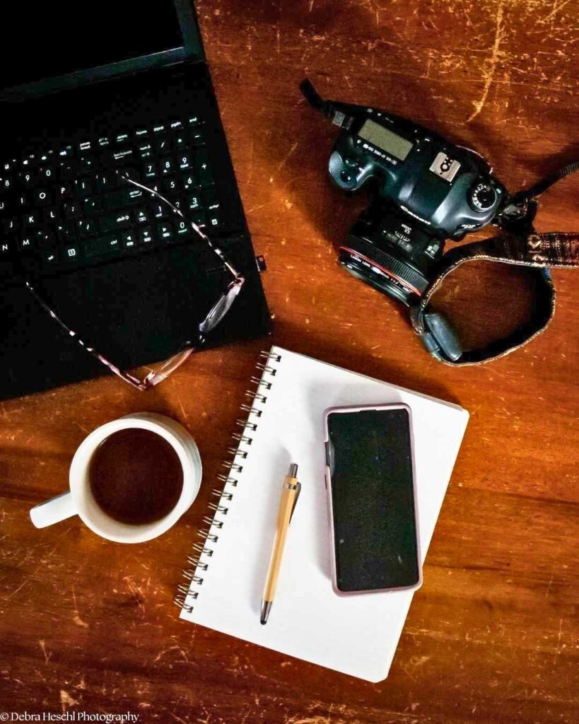 Overhead view of a photographer's workspace with a Canon DSLR camera, laptop, glasses, coffee cup, notebook, pen, and phone on a wooden desk.