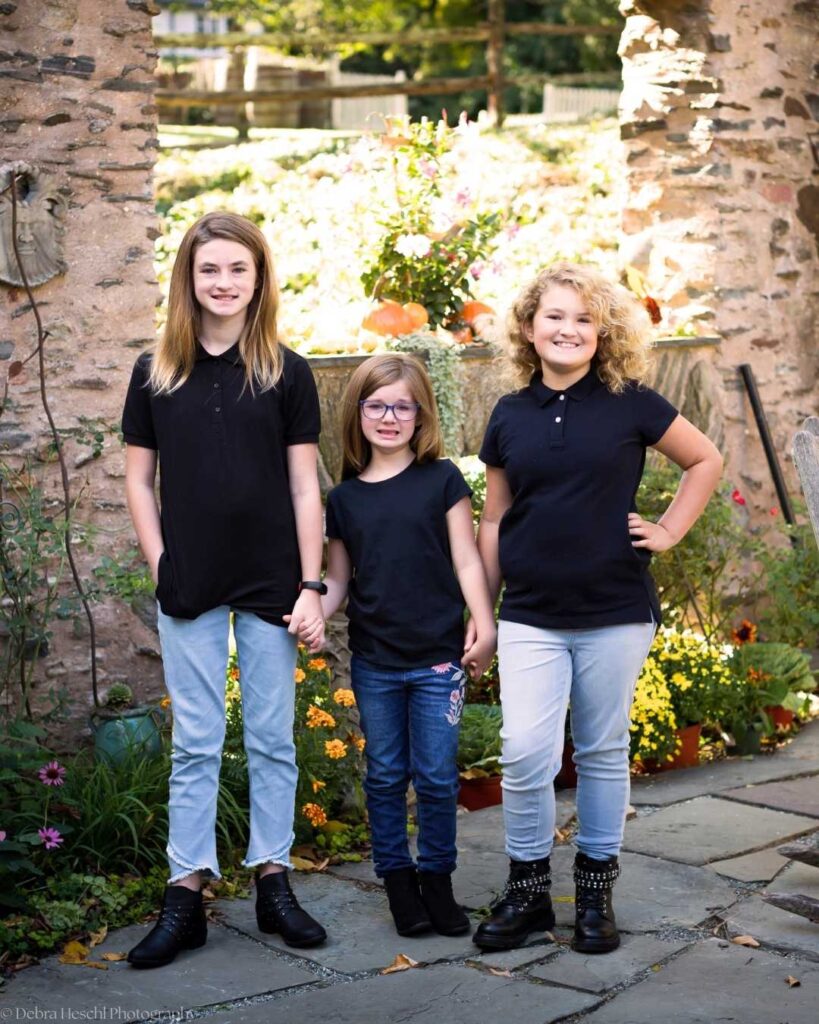 Three sisters holding hands and smiling during an outdoor family photography session at Manor House of Prophecy Creek in Blue Bell, Pennsylvania.