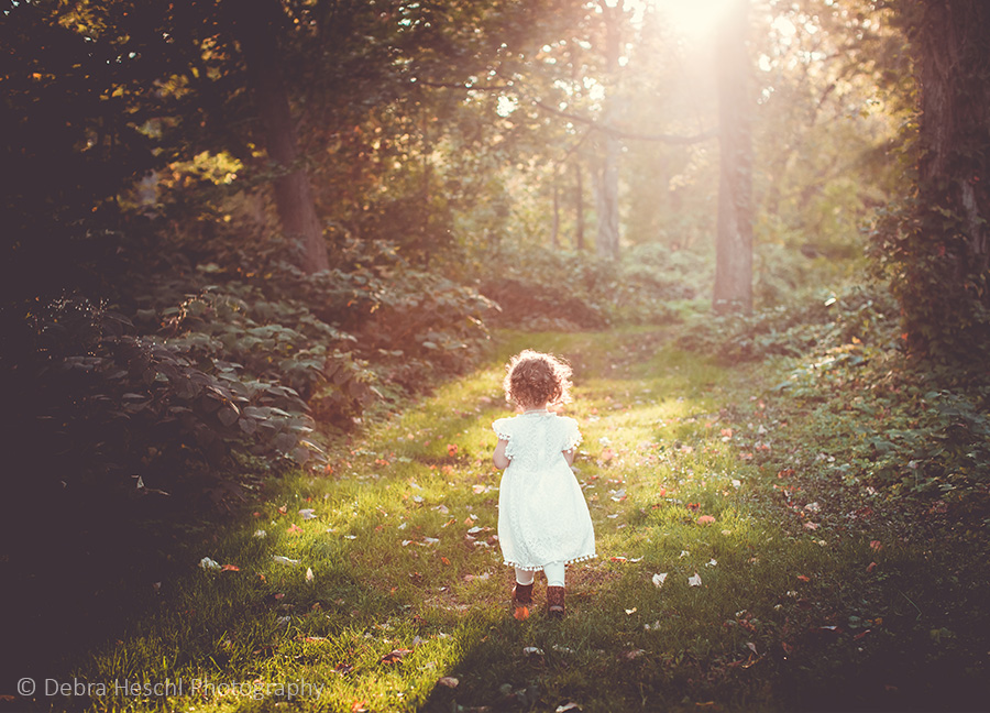 Toddler girl in a white dress walking down a sunlit wooded path at Harriton House in Bryn Mawr, PA