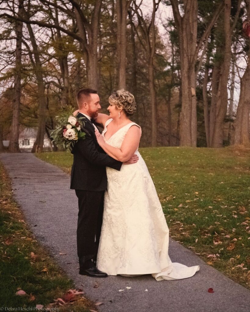 Bride and groom embracing on a paved park path surrounded by tall trees and fallen leaves at an autumn wedding