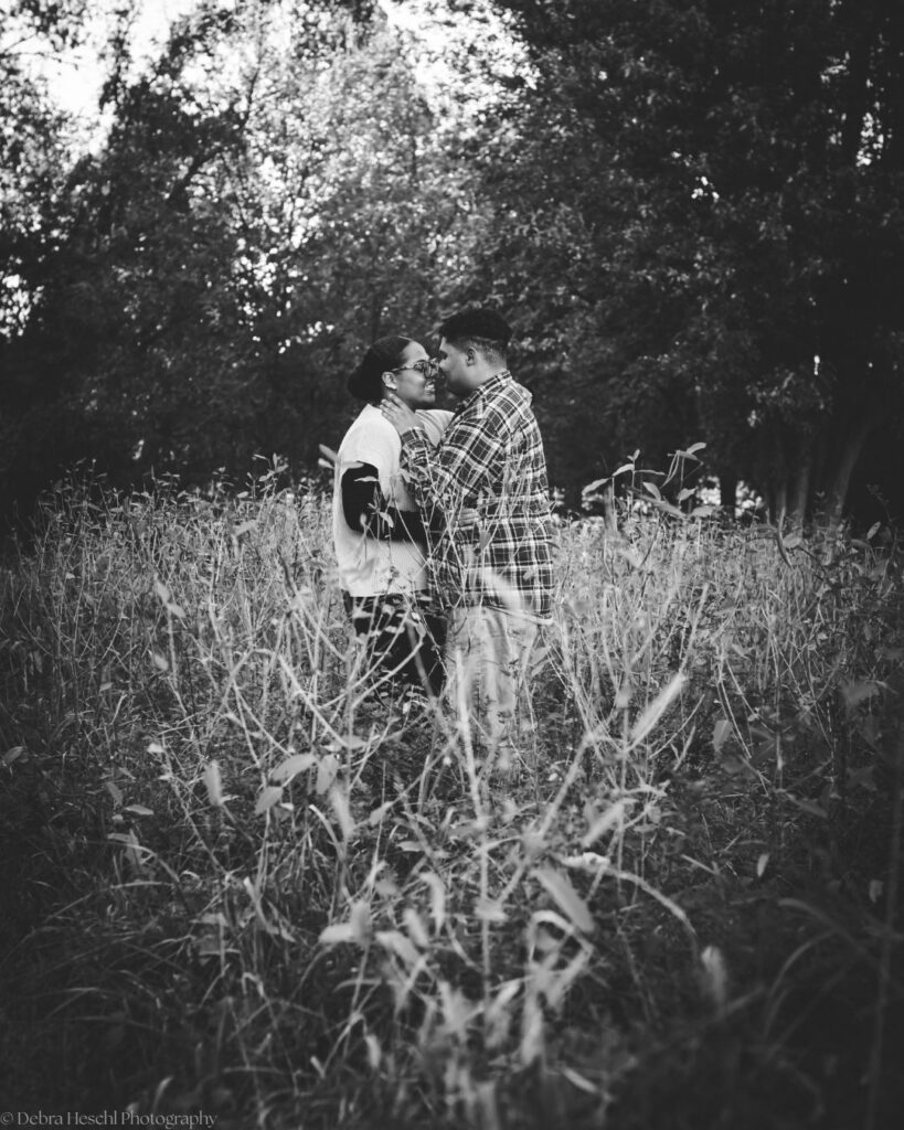 Black and white photo of Hannah and Richard embracing nose to nose in tall grass with trees behind them at Manor House.