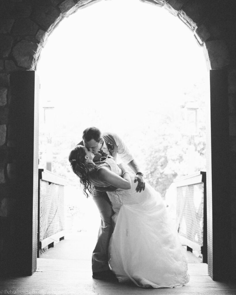 Black and white photo of a groom dipping his bride for a kiss beneath a stone archway at the start of a wooden bridge