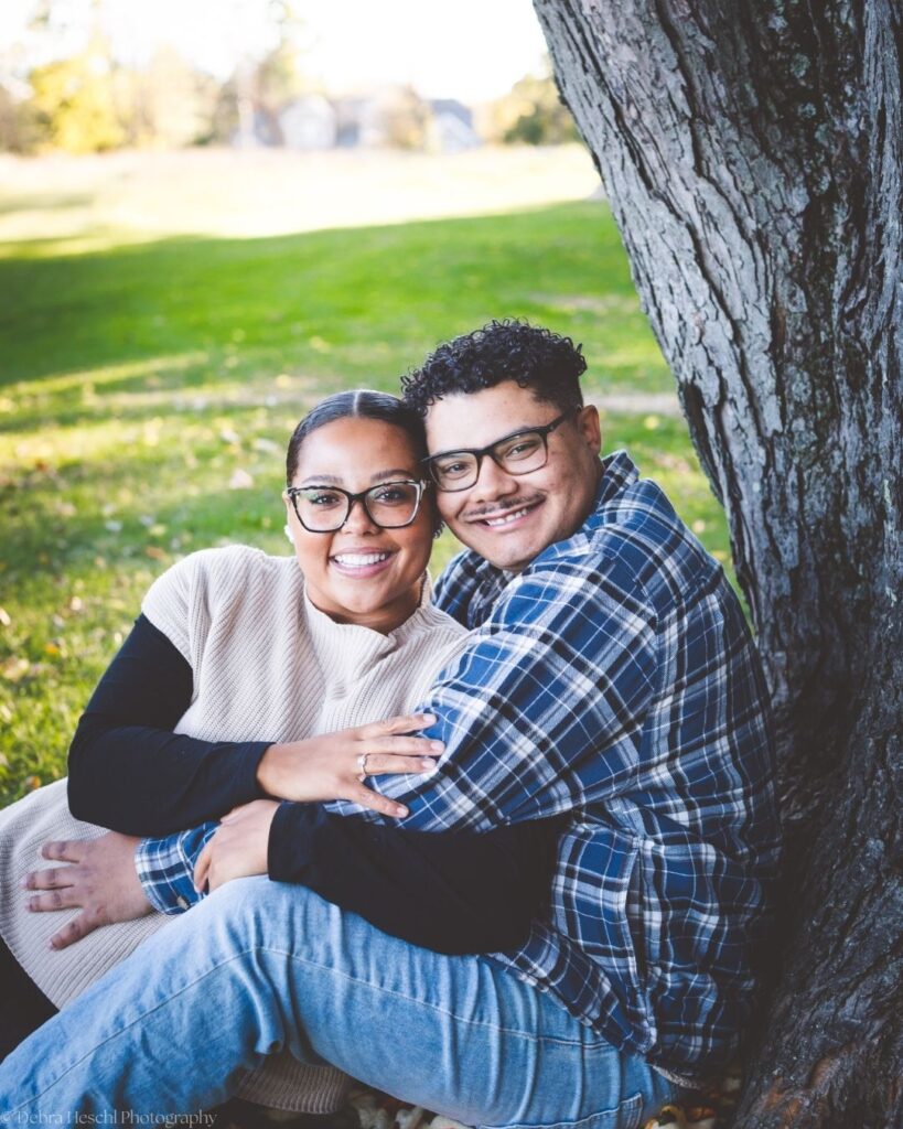 Hannah and Richard smiling and snuggling together at the base of a large tree on the grass at Manor House.