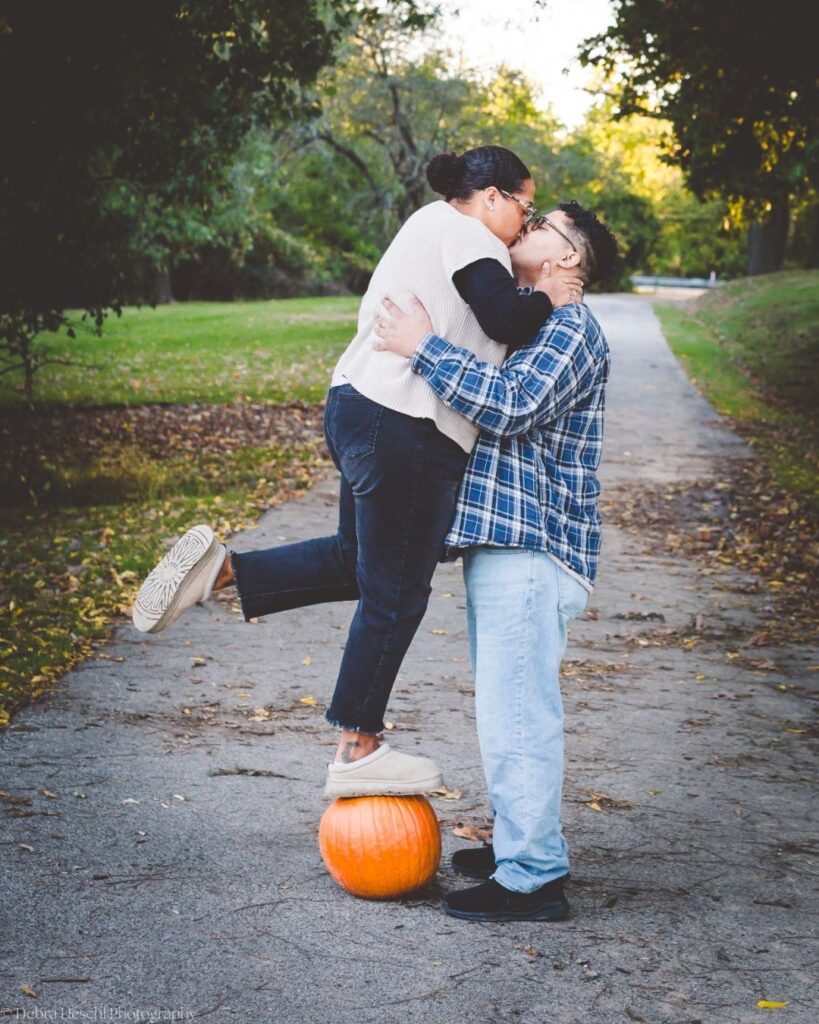 Hannah and Richard sharing a playful kiss on a Manor House path, with Hannah's foot popped up on an orange pumpkin.