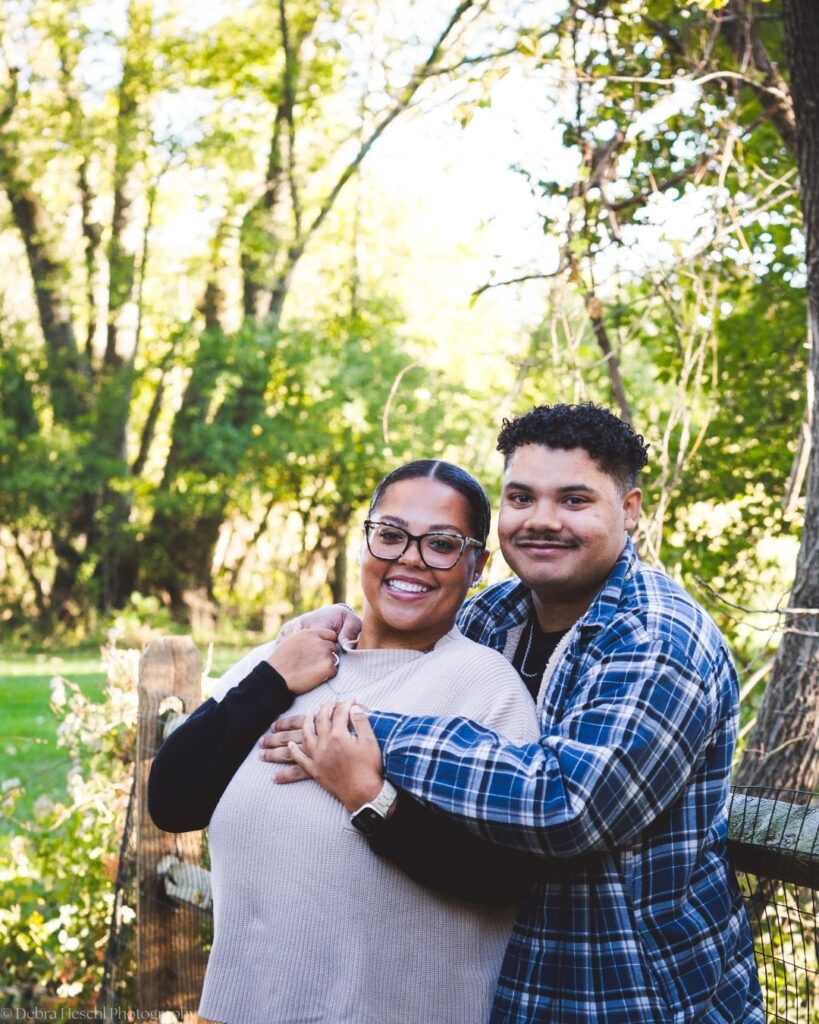 Hannah and Richard smiling and hugging beside a wooden fence surrounded by green trees during their fall photo session.