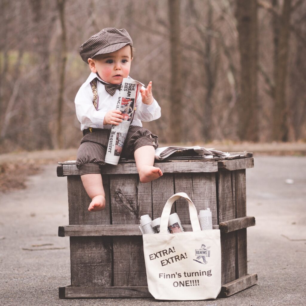 Toddler boy in a newsboy cap, bow tie, and suspenders sitting on a wooden crate beside an “Extra! Extra! Finn’s turning one” tote bag, a vintage newspaper-themed first birthday portrait in Bridgeport, PA