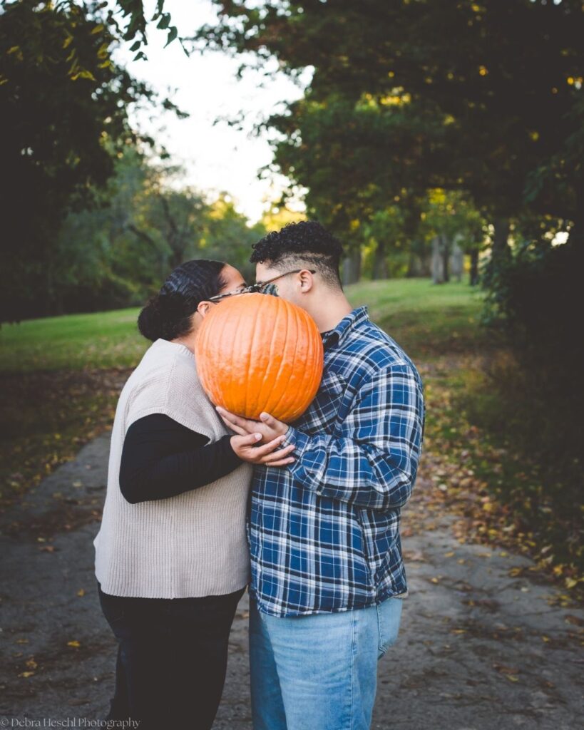 Hannah and Richard kissing behind a large orange pumpkin on a leaf-covered path during their fall photo session at Manor House.