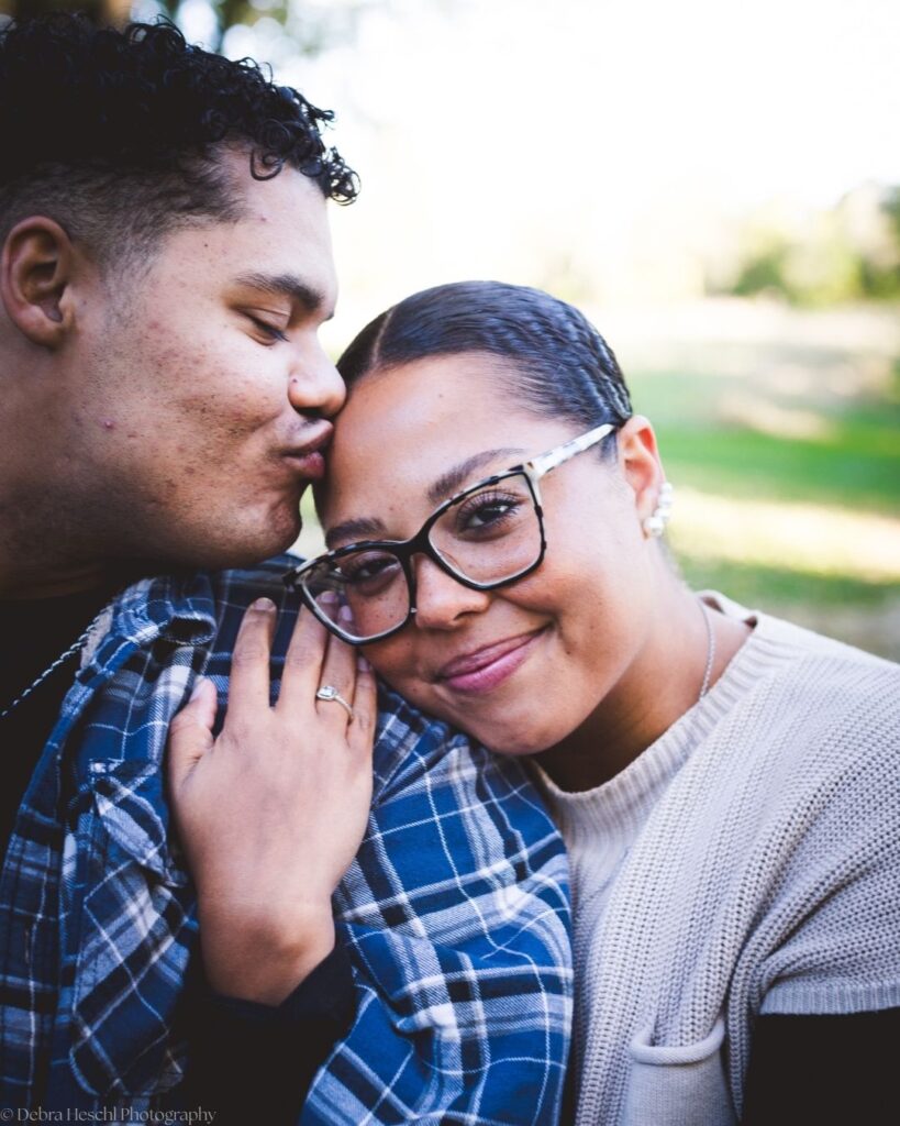Close-up of Richard kissing Hannah's forehead while she smiles at the camera, with a soft green park behind them.