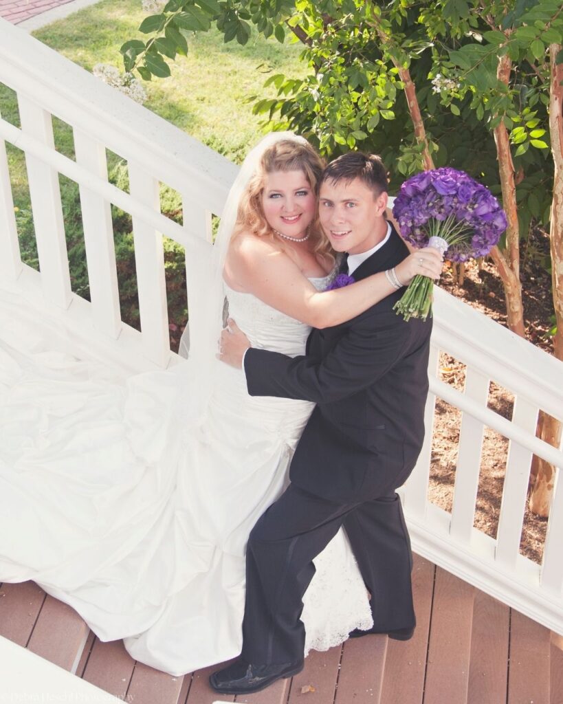 Smiling bride and groom embracing on a white wooden staircase, shot from above, with the bride holding a purple rose bouquet