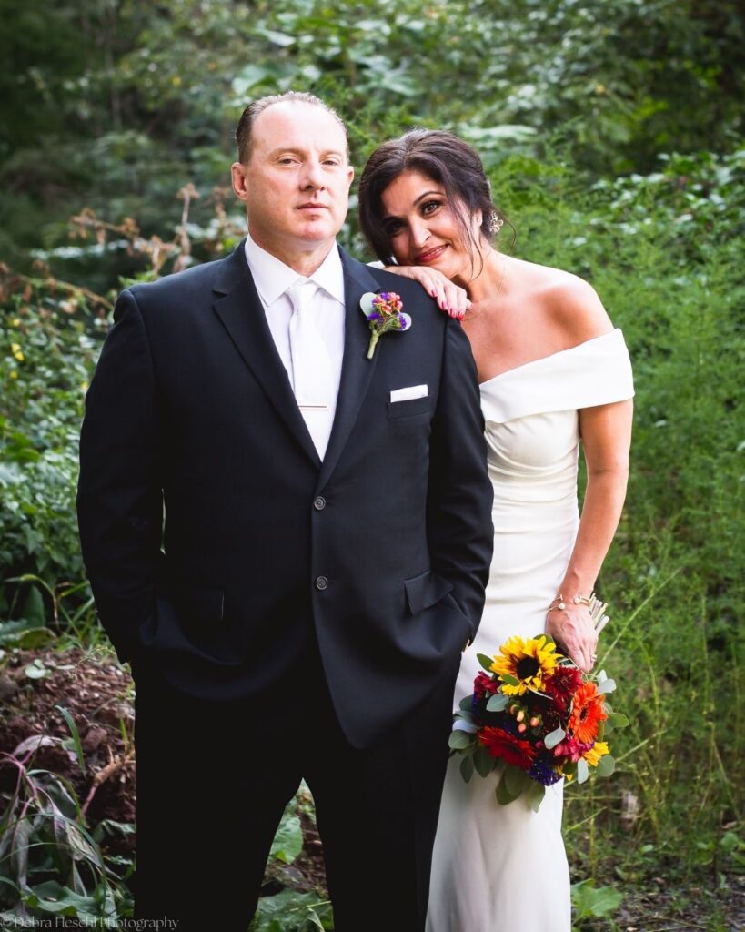 Bride in an off-the-shoulder gown leaning on her groom's shoulder, holding a sunflower bouquet in a lush green garden